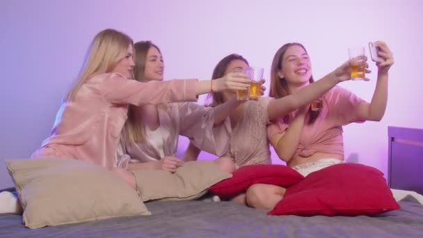 Vier gelukkige mooie jonge vrouwen in pyjama en met bril zit op bed en opnemen video felicitatie - Video