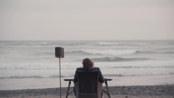 Medium back-view shot of unrecognizable man sitting at desk in front of blue ocean on isolated beach - Footage, Video