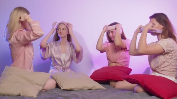 Vier gelukkige mooie jonge vrouwen in pyjama zit op bed en zet hoofdbanden op vrijgezellenfeest - Video