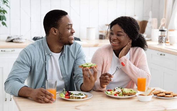 Positieve zwarte echtgenoten eten smakelijk ontbijt en drinken sinaasappelsap in de keuken - Foto, afbeelding