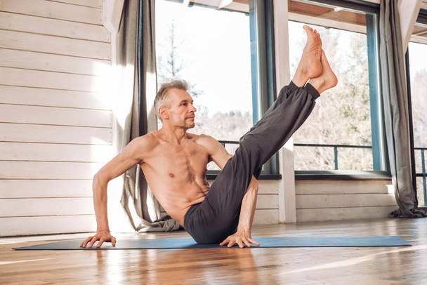 Caucasian senior man practicing yoga classic asana in studio, training in front of a window. Copy, empty space for text. - Фото, изображение