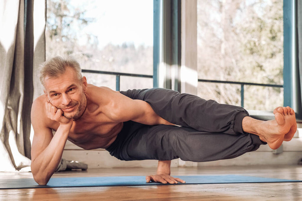 Senior man with a naked torso practicing yoga classic asana training in studio in front of a window. Copy, empty space for text. - Foto, afbeelding