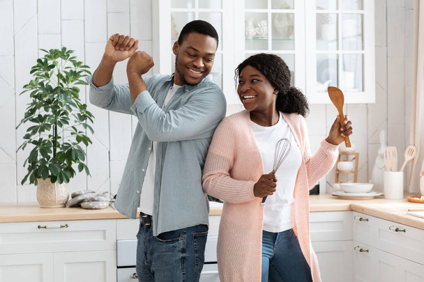 Ευτυχισμένο ζευγάρι Αφρικάνων που διασκεδάζουν στην κουζίνα, χορεύουν με τα ούτεκ στα χέρια - Φωτογραφία, εικόνα