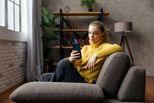 Cheerful young woman using mobile phone while sitting on a sofa at home. - Photo, image