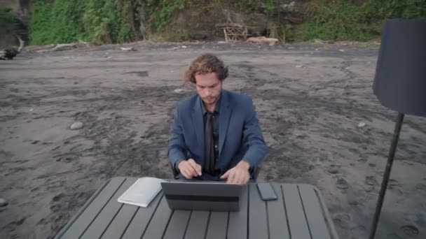 Obloukový dron záběr obchodníka v obleku sedí za stolem s laptopem na prázdném pobřeží oceánu za denního světla - Záběry, video