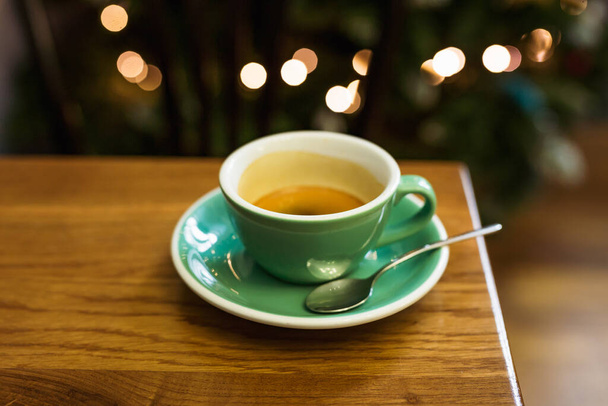 Una foto de estilo de vida horizontal de una taza verde de café negro en un platillo, una cuchara, luces bokeh en el fondo, enfoque selectivo - Foto, imagen
