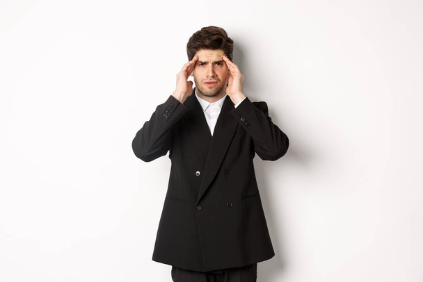 Image d'un homme d'affaires en costume noir, touchant la tête et ayant l'air étourdi, ressentant des maux de tête douloureux, debout sur un fond blanc - Photo, image