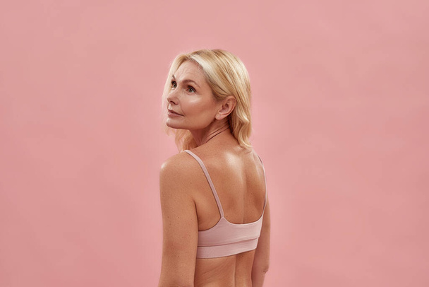 Aging process. Rear view of a beautiful middle aged caucasian blonde woman wearing underwear looking away while posing isolated over pink background - Fotó, kép
