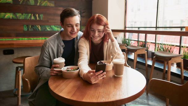 Happy teenage couple smiling while taking selfies using smartphone, sitting in a cafe together on a daytime - Фото, изображение