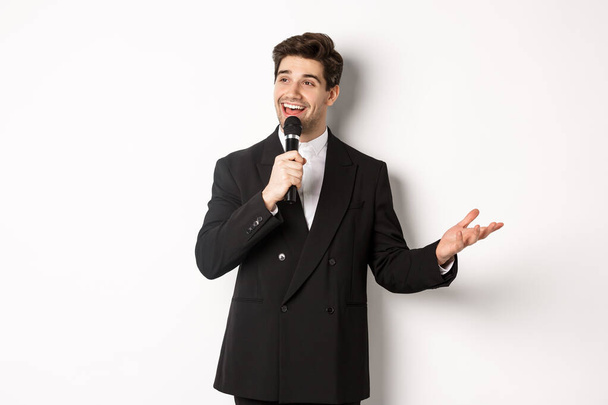 Portret van een knappe man in zwart pak die een lied zingt, een microfoon vasthoudt en een toespraak houdt, staande tegen een witte achtergrond - Foto, afbeelding