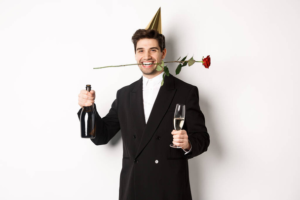 Забавный парень в модном костюме, празднует и устраивает вечеринку, держит розу в зубах и шампанское, стоит на белом фоне - Фото, изображение