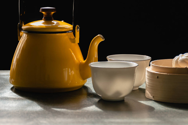 Two white cups and yellow pot of tea or coffee, Steamed buns in wooden basket, Morning tea on black background, Food that provides warmth to body and has health benefits in winter season. - Fotó, kép