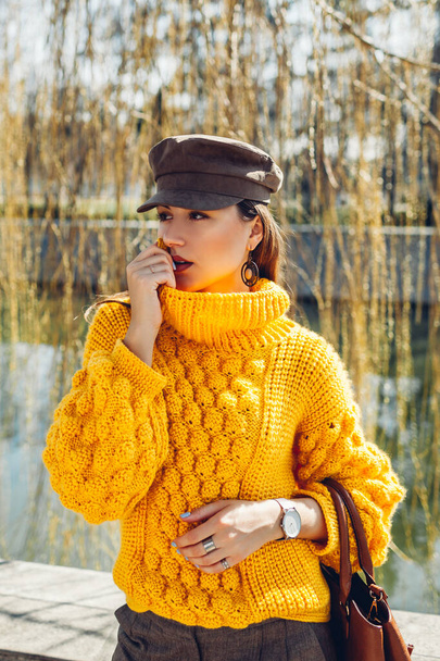 Fiatal nő visel elegáns sárga pulóver kalap és tartja kézitáska pihentető parkban. Tavaszi női ruhák és kiegészítők. Divatot. 2021-es szín - Fotó, kép