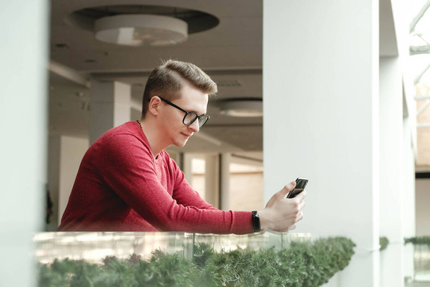 Πορτρέτο ενός νεαρού άνδρα με γυαλιά και με ένα smartphone στα χέρια του κοντά σε ένα γυάλινο φράχτη στο εμπορικό κέντρο. Έννοια επικοινωνίας. - Φωτογραφία, εικόνα