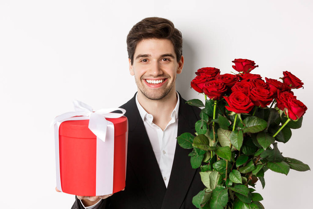 Close-up of handsome bearded man in suit, holding present and bouquet of red roses, smiling at camera, standing against white background - Photo, Image