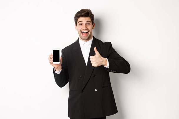 Εικόνα ενός όμορφου άνδρα επιχειρηματία με μαύρο κοστούμι, που συνιστά εφαρμογή ή ηλεκτρονικό κατάστημα, που δείχνει τους αντίχειρες προς τα πάνω και οθόνη smartphone, που στέκεται πάνω από λευκό φόντο - Φωτογραφία, εικόνα