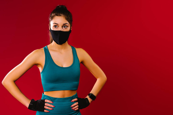 Portrait of a sportive girl in a sport top and a black protective mask on a red background with blank space for advertising. The concept of sports during a pandemic. - Foto, imagen