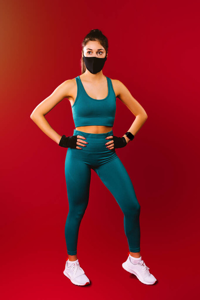 Full-length photo. A pumped-up girl in sportswear and a black medical protective mask on a red background. The concept of sports during a pandemic. - Fotoğraf, Görsel