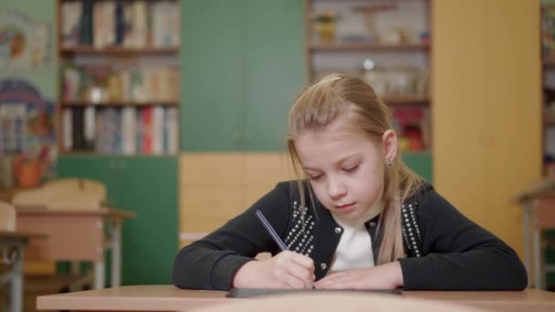 A school-age girl sits at a desk in a school class. She is busy with a study assignment. The concept of education in a school led by teachers. - Footage, Video