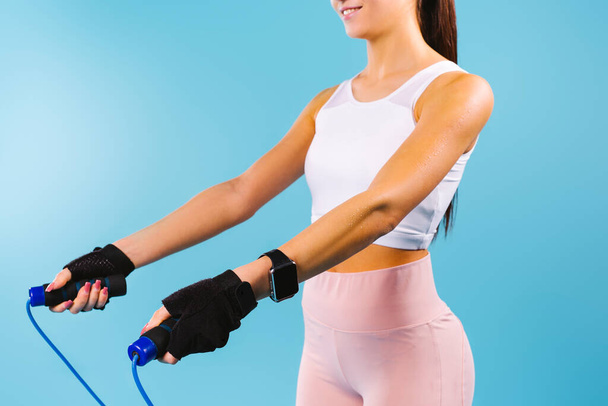Cropped photo of a caucasian athlete girl in a sportwear top and a blue skipping rope, workout on a blue background. - Foto, Imagem