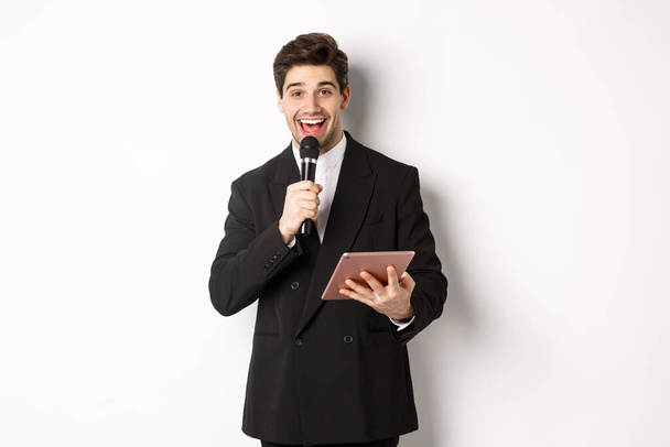 Image of handsome male performer, giving a speech, entertain people at party, holding microphone and digital tablet, standing over white background - Photo, Image