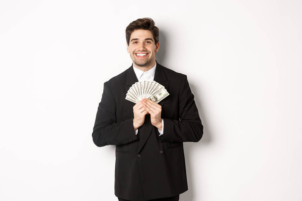 Portré boldog és elégedett jóképű férfi öltönyben, ölelés pénzt, és úgy néz ki, elégedett, felett álló fehér háttér - Fotó, kép
