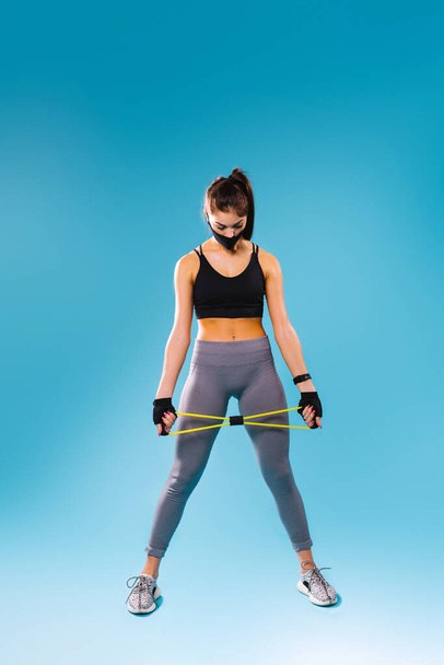 A full-length photo of a pumped-up girl in a protective black mask and with a resitance band in her hands. Looks down on a blue background with blank space. - Foto, Imagen