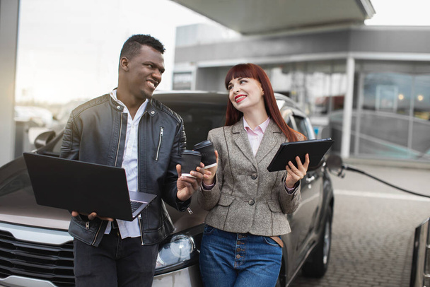 Business couple, Caucasian woman and African man, looking each other with smile, standing together outdoors near the car during the coffee break, refueling auto at gas station - Photo, image