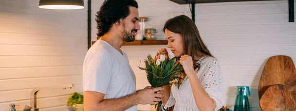 Internationale Vrouwendag. Jong stel in de ochtend in de keuken thuis. Een man geeft een vrouw een boeket tulpen op 8 maart in een mooie stijlvolle keuken. - Foto, afbeelding