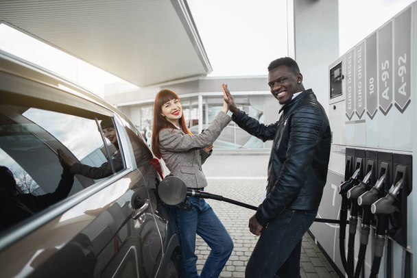 Couple at gas station. Happy joyful multiracial couple of friends or colleagues, posing to camera, having fun while refueling luxury car at petrol station, giving high five each other and smiling - Foto, imagen