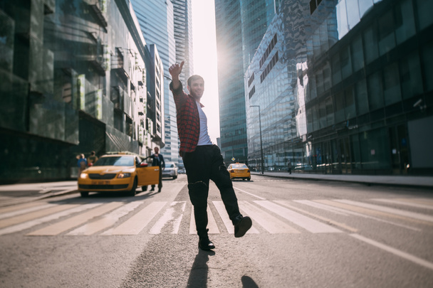 Un hombre posa en un paso de peatones en el centro de negocios de la ciudad. Joven chico elegante posando al atardecer en una gran ciudad en el fondo de los rascacielos - Foto, imagen