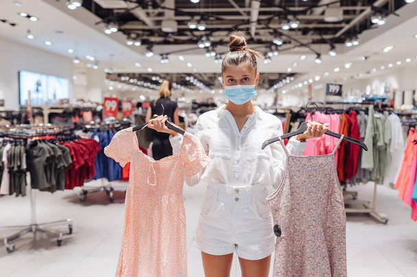 Jonge vrouw winkelen kleding in kleding boetiek met beschermende gezichtsmasker - Foto, afbeelding