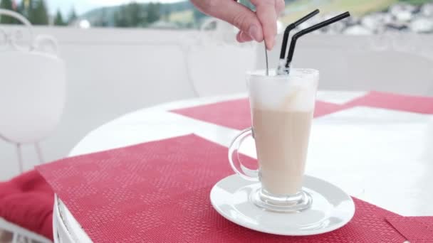 Weiße Hand mit einem Teelöffel, der in einer großen Kaffeetasse köstlichen Latte rührt. Zwei Plastikhalme in einem Getränk. Ein entspannter Morgen im Freien mit Kaffee auf einem Wochenendausflug. Einmalstroh im Glas. - Filmmaterial, Video