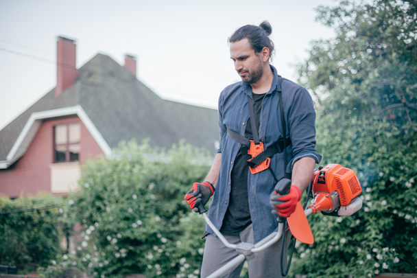 Ένας άντρας κουρεύει το γρασίδι με μια μηχανή χειρός στον κήπο. Ο νεαρός φροντίζει το γκαζόν και δουλεύει στον κήπο. - Φωτογραφία, εικόνα