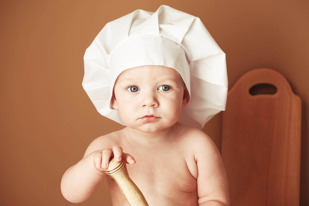 Portrait little baby boy in a chef's hat sitting on the table holds a wooden rolling pin on a brown background. Copy, empty space for text. - Foto, Bild