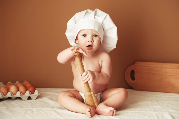 Malý chlapeček v kuchařském klobouku, který sedí na stole, drží dřevěný váleček na hnědém pozadí. Kopírovat, prázdné místo pro text. - Fotografie, Obrázek