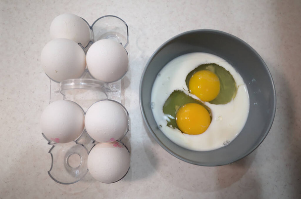 Dos huevos crudos en un bol con leche cerca de huevos blancos en cáscara de huevo en bandeja de huevo. Ingredientes alimentarios para cocinar tortilla para el desayuno - Foto, Imagen