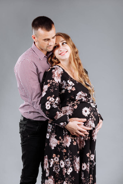 Una mujer embarazada y su marido se abrazan sobre un fondo gris en el estudio - Foto, Imagen