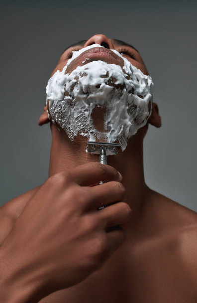 Close up of young african american man with foam applied on his face using steel razor for removing stubble from neck isolated over gray background - Foto, Bild
