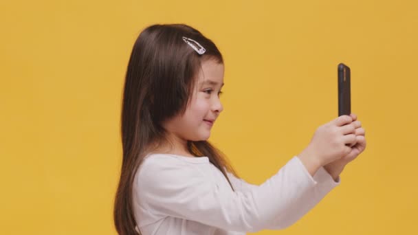 Generación Z. Adorable niña haciendo selfie en la cámara del teléfono inteligente, sonriendo y mostrando la lengua hacia fuera, fondo naranja - Metraje, vídeo
