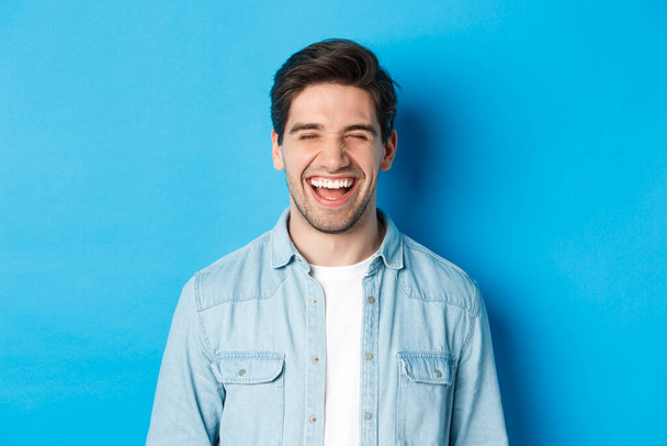 Крупним планом щасливий молодий бородатий чоловік в повсякденному одязі, сміючись і відчуваючи радість, стоїть над синім тлом
 - Фото, зображення