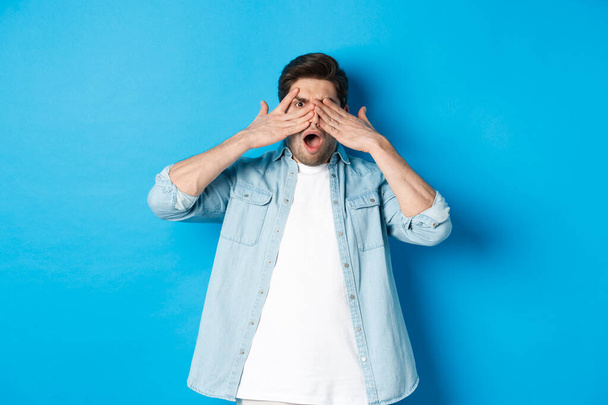 Shocked man covering eyes and peeking through fingers, stare at something embarrassing, standing against blue background - Photo, Image
