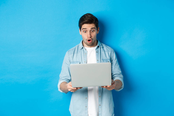 Imponujący biały facet patrząc na ekranie laptopa ze zdumieniem, sprawdzając promo w Internecie, stojąc na niebieskim tle - Zdjęcie, obraz