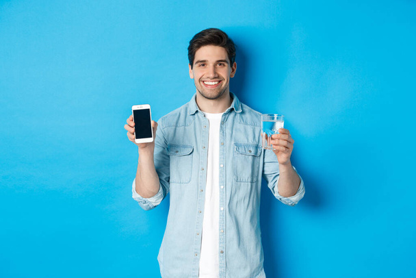 Nuori mies hallita veden tasapaino älypuhelin app, osoittaa mobiili näyttö sovellus ja hymyilevä, seisoo sininen tausta - Valokuva, kuva
