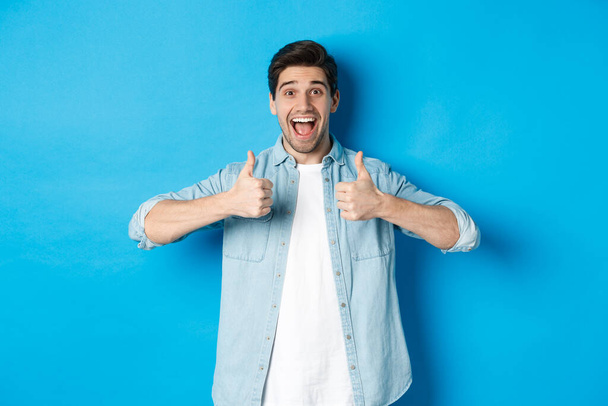 Smiling adult man showing thumbs up with excited face, like something awesome, approving product, standing against blue background - Photo, Image