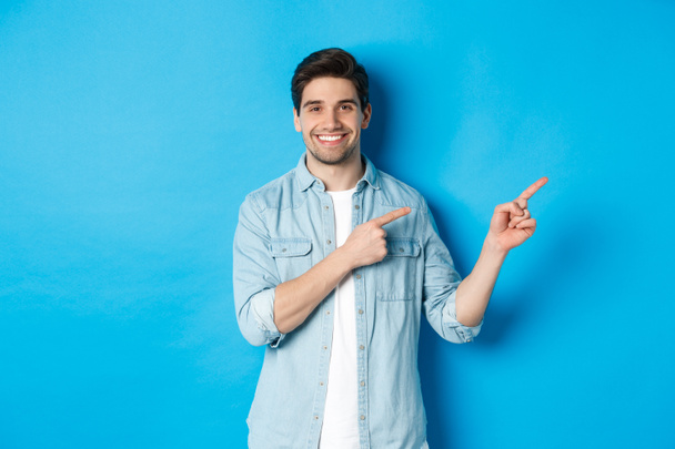 Πορτρέτο του ελκυστικού ενήλικα χαμογελώντας, δείχνοντας τα δάχτυλα δεξιά στο λογότυπο ή πανό, δείχνει διαφήμιση σε μπλε φόντο - Φωτογραφία, εικόνα