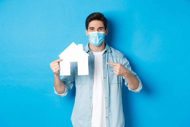 Concept of coronavirus, quarantine and social distancing. Young man searching aparment for rent, business loans, pointing at house model, wearing medical mask, blue background - Photo, Image