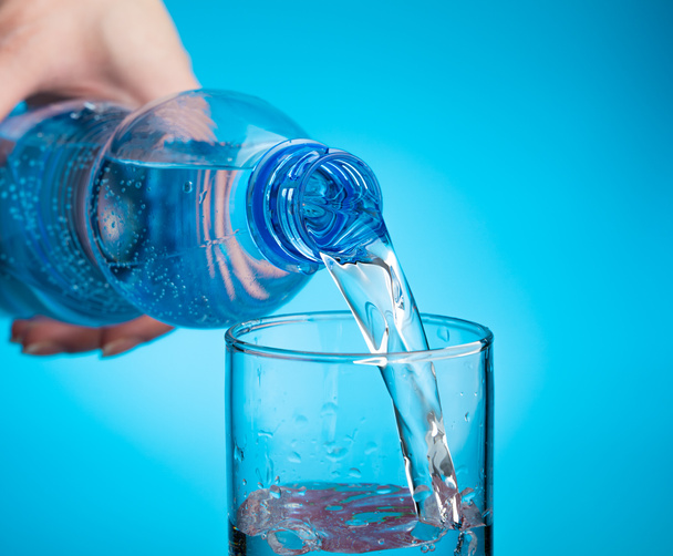 Water in een glas gieten op een lichtblauwe achtergrond. Close-up, zijaanzicht. - Foto, afbeelding