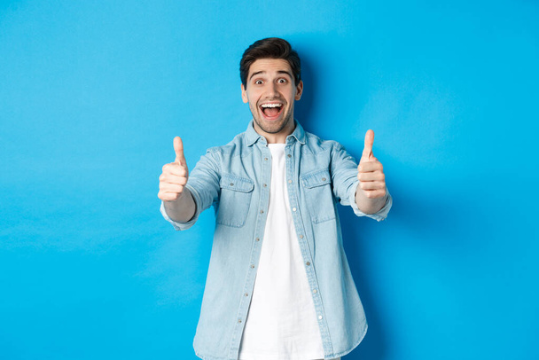 Usmívající se sebevědomý muž ukazuje palce nahoru s vzrušeným obličejem, jako něco úžasného, schvalující produkt, stojí na modrém pozadí - Fotografie, Obrázek