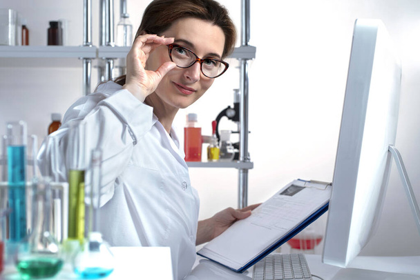 Aktywna kobieta naukowiec koryguje okulary sprawdzając dane w notatniku papieru w laboratorium chemicznym. Przyjacielska twarz. Scena plenerowa stylu życia - Zdjęcie, obraz